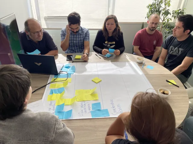 Equipo de EMOJ alrededor de una mesa, trabajando con notas adhesivas sobre un papelógrafo.