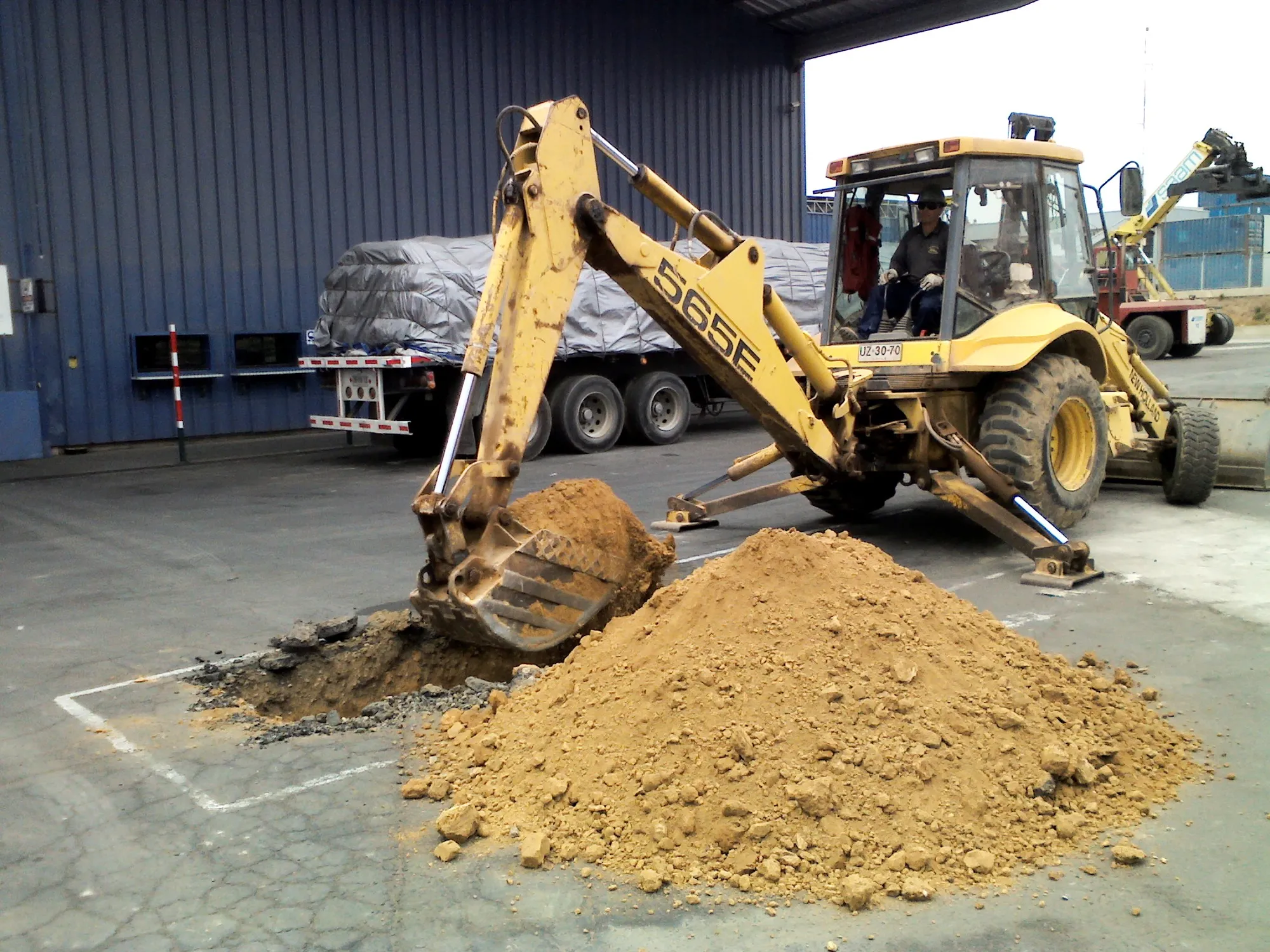 Retroexcavadora abriendo una calicata junto a un acopio de tierra en un recinto industrial.