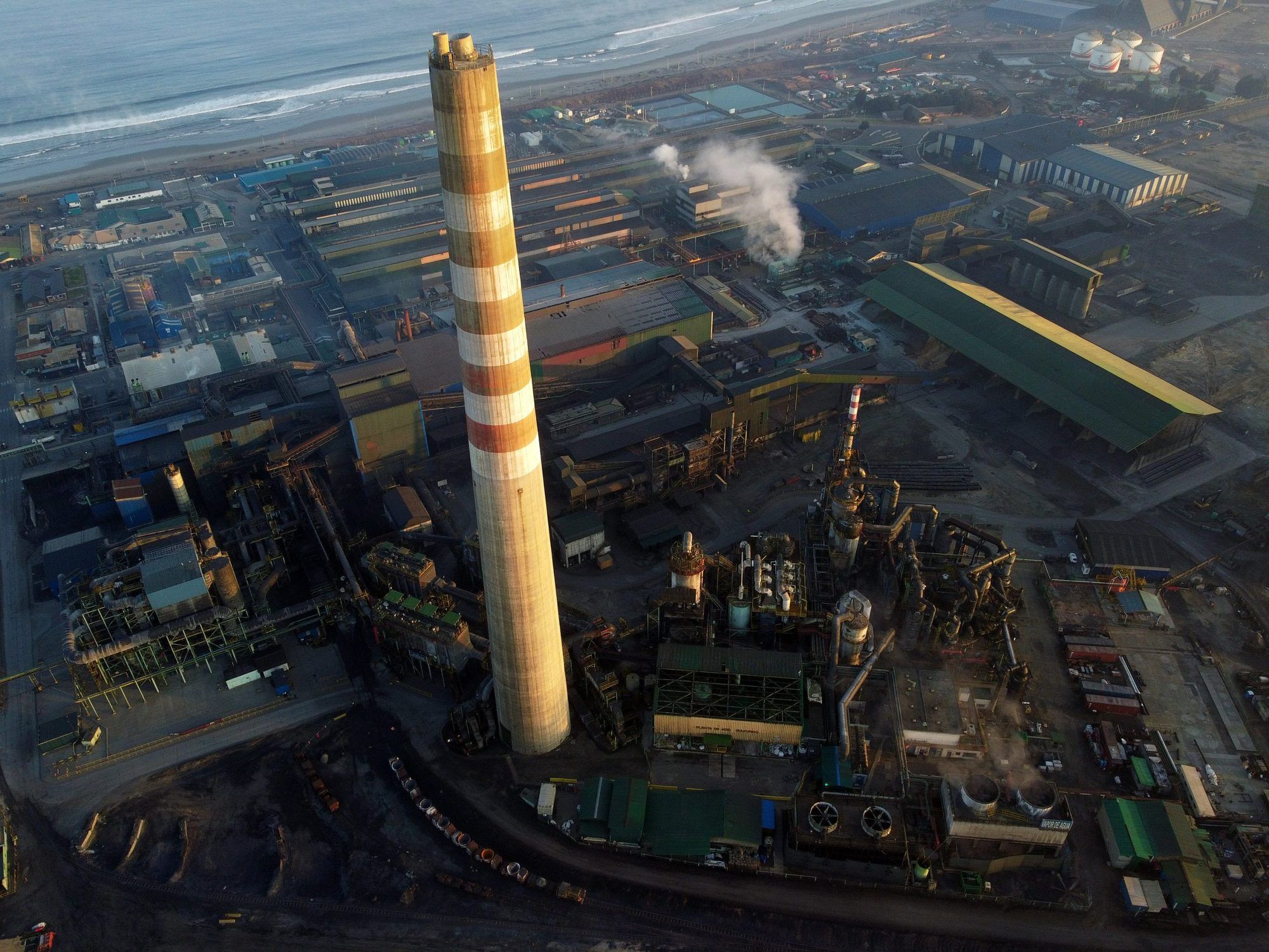 Vista aérea de un complejo industrial costero con una chimenea alta.